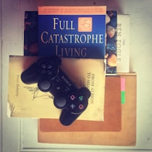 A black gaming controller is resting on top of a stack of books. The books include titles such as 'Full Catastrophe Living,' 'From Albion to Shangri-La,' and 'The Ben Fogle.' The overall setting suggests a blend of leisure and intellectual pursuits.