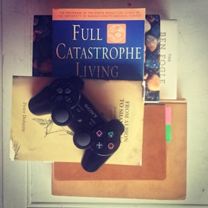 A black gaming controller is resting on top of a stack of books. The books include titles such as 'Full Catastrophe Living,' 'From Albion to Shangri-La,' and 'The Ben Fogle.' The overall setting suggests a blend of leisure and intellectual pursuits.