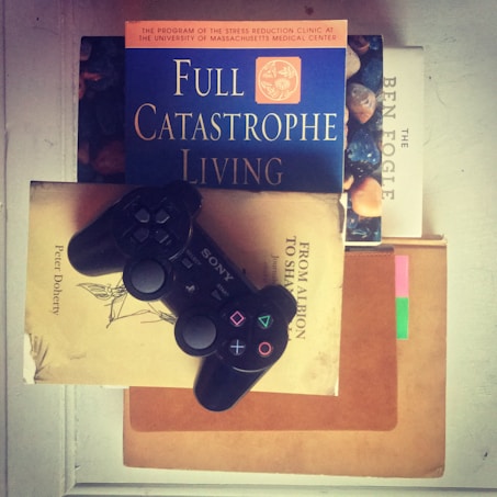 A black gaming controller is resting on top of a stack of books. The books include titles such as 'Full Catastrophe Living,' 'From Albion to Shangri-La,' and 'The Ben Fogle.' The overall setting suggests a blend of leisure and intellectual pursuits.