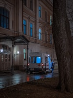 An ambulance with illuminated lights is parked outside a building at night. A person wearing blue protective gear stands nearby on a wet pavement next to bare trees.