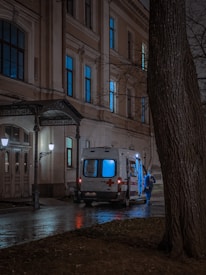 An ambulance with illuminated lights is parked outside a building at night. A person wearing blue protective gear stands nearby on a wet pavement next to bare trees.