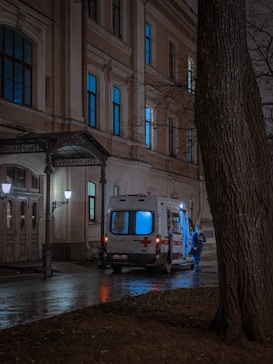 An ambulance with illuminated lights is parked outside a building at night. A person wearing blue protective gear stands nearby on a wet pavement next to bare trees.