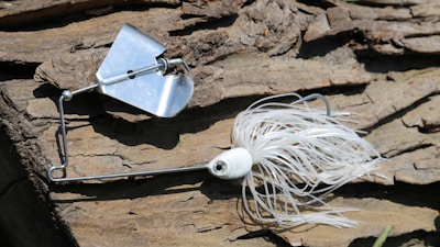 A white and silver fishing lure rests on a textured wooden surface. The lure has a metal spinner and fringed skirt, designed to attract fish. Sunlight casts shadows on the wood, highlighting its rugged texture.