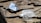 A white and silver fishing lure rests on a textured wooden surface. The lure has a metal spinner and fringed skirt, designed to attract fish. Sunlight casts shadows on the wood, highlighting its rugged texture.