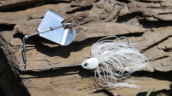 A white and silver fishing lure rests on a textured wooden surface. The lure has a metal spinner and fringed skirt, designed to attract fish. Sunlight casts shadows on the wood, highlighting its rugged texture.