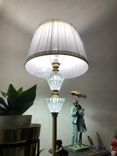 A decorative table lamp with a large, ornate shade stands prominently alongside a small potted plant with green leaves. Below the lamp, there is a detailed sculpture of a couple intimately embracing under an umbrella, adding an artistic and romantic element to the scene.