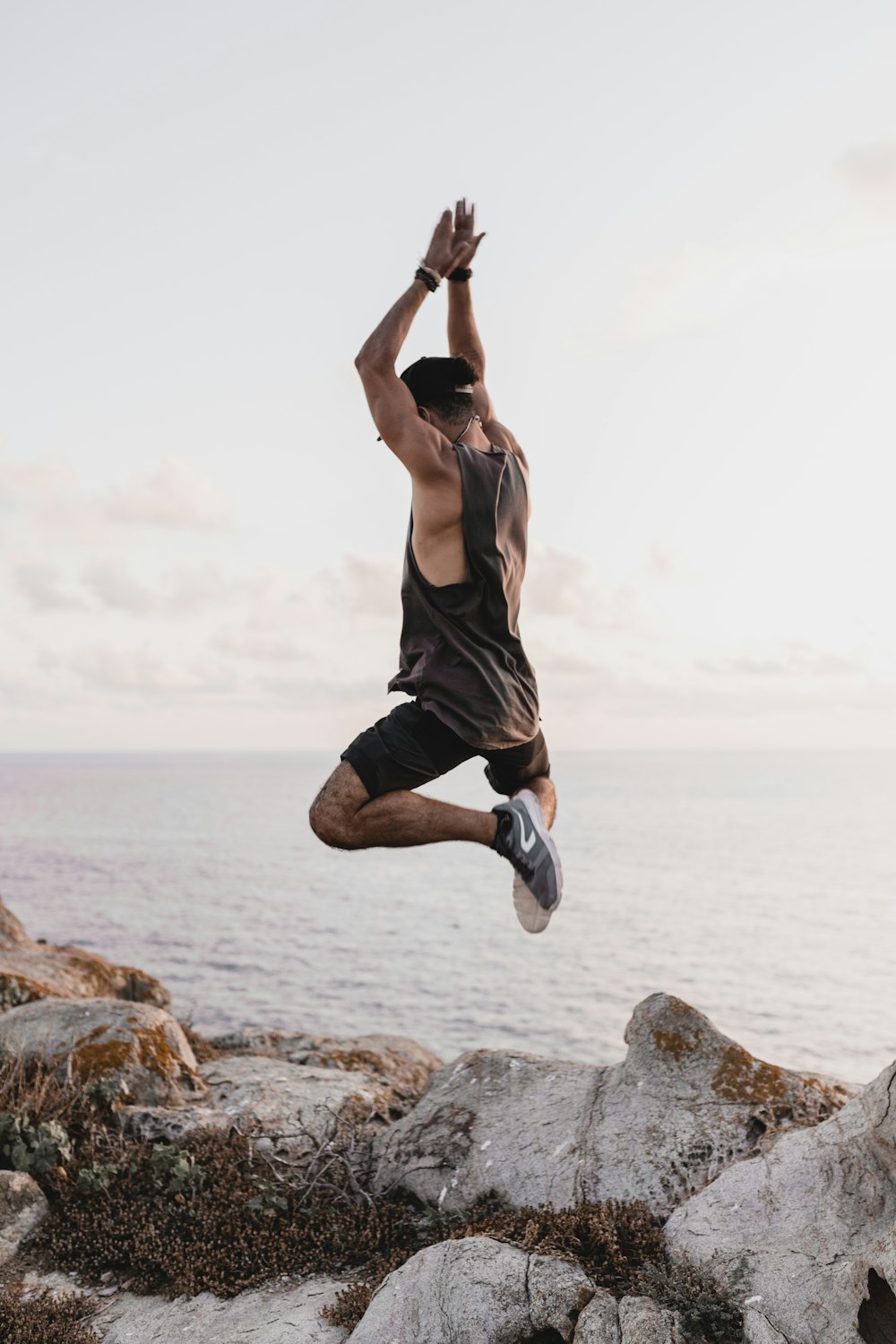 woman in black tank top and black shorts jumping on brown rock near sea during daytime