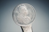An antique silver coin with intricate engravings, showing a profile portrait in relief. Surrounding the portrait are inscriptions in a circular pattern.