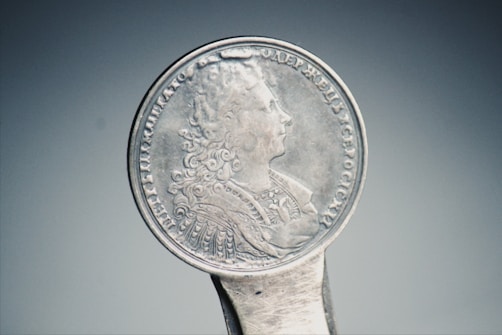 An antique silver coin with intricate engravings, showing a profile portrait in relief. Surrounding the portrait are inscriptions in a circular pattern.
