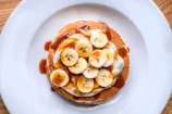Stack of fluffy pancakes drizzled with maple syrup and topped with sliced bananas and walnuts.
