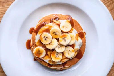 Stack of fluffy pancakes drizzled with maple syrup and topped with sliced bananas and walnuts.