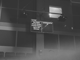 A black and white image featuring a screen displaying text in a dimly lit room. The content on the screen includes phrases like 'Way Maker' and 'Light in the darkness', as well as a timestamp of 7:11 AM. The screen is mounted high on a wall with visible speakers and ceiling lights.