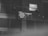A black and white image featuring a screen displaying text in a dimly lit room. The content on the screen includes phrases like 'Way Maker' and 'Light in the darkness', as well as a timestamp of 7:11 AM. The screen is mounted high on a wall with visible speakers and ceiling lights.