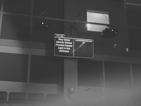 A black and white image featuring a screen displaying text in a dimly lit room. The content on the screen includes phrases like 'Way Maker' and 'Light in the darkness', as well as a timestamp of 7:11 AM. The screen is mounted high on a wall with visible speakers and ceiling lights.