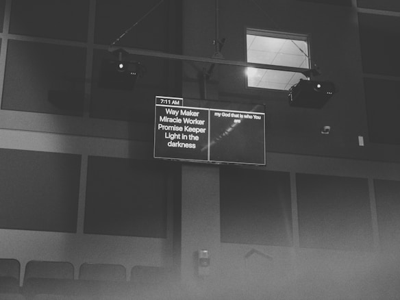 A black and white image featuring a screen displaying text in a dimly lit room. The content on the screen includes phrases like 'Way Maker' and 'Light in the darkness', as well as a timestamp of 7:11 AM. The screen is mounted high on a wall with visible speakers and ceiling lights.