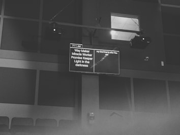 A black and white image featuring a screen displaying text in a dimly lit room. The content on the screen includes phrases like 'Way Maker' and 'Light in the darkness', as well as a timestamp of 7:11 AM. The screen is mounted high on a wall with visible speakers and ceiling lights.