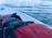 A close-up of a red life jacket lying on a paddleboard, with calm, reflective water visible in the background. The jacket is partially wet and features a strap and buckle.
