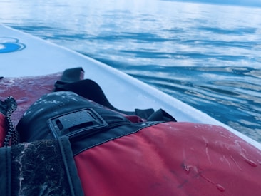 A close-up of a red life jacket lying on a paddleboard, with calm, reflective water visible in the background. The jacket is partially wet and features a strap and buckle.