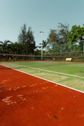 An outdoor tennis court with a red and green playing surface surrounded by a fence and trees in the background. Two tennis balls are placed near the net, and there is a stack of yellow tennis ball containers near the fence.