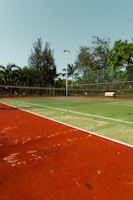An outdoor tennis court with a red and green playing surface surrounded by a fence and trees in the background. Two tennis balls are placed near the net, and there is a stack of yellow tennis ball containers near the fence.