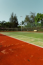 An outdoor tennis court with a red and green playing surface surrounded by a fence and trees in the background. Two tennis balls are placed near the net, and there is a stack of yellow tennis ball containers near the fence.