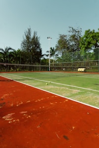 An outdoor tennis court with a red and green playing surface surrounded by a fence and trees in the background. Two tennis balls are placed near the net, and there is a stack of yellow tennis ball containers near the fence.