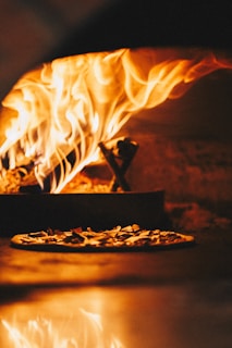 Wood-fired oven with bright flames engulfing the wood, reflecting a warm and fiery atmosphere. A pizza topped with ingredients is seen in the foreground, baking inside the oven.