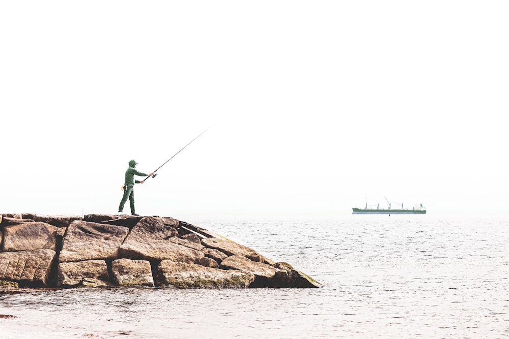 man in green shirt and black pants standing on brown rock near body of water during