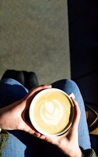 A person holds a large white mug filled with a hot beverage featuring latte art in the shape of a heart and leaf pattern. The person is wearing blue jeans, and their hands are softly cradling the mug.