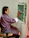 A person is seated and painting an artwork on a canvas attached to the wall. The artwork features a colorful tree with leaves and flowers in a natural setting. The person is wearing a traditional striped garment and is focused on the painting.