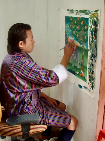 A person is seated and painting an artwork on a canvas attached to the wall. The artwork features a colorful tree with leaves and flowers in a natural setting. The person is wearing a traditional striped garment and is focused on the painting.