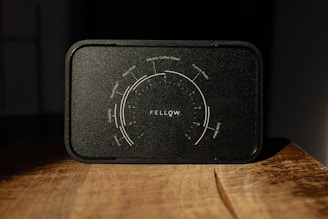 A black coffee brewing guide from Fellow, featuring a detailed dial with various brewing methods including pour-over, electric coffee maker, French press, Aeropress, and cold brew. The guide is positioned on a wooden surface, lit dramatically with shadows enhancing its texture.