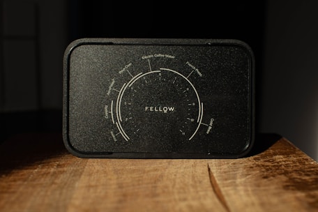 A black coffee brewing guide from Fellow, featuring a detailed dial with various brewing methods including pour-over, electric coffee maker, French press, Aeropress, and cold brew. The guide is positioned on a wooden surface, lit dramatically with shadows enhancing its texture.
