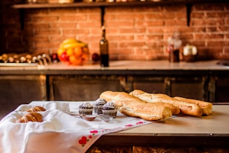 A kitchen counter displays a cloth-covered table with an assortment of baked goods including croissants, chocolate muffins dusted with powdered sugar, and several baguettes. In the background, a rustic kitchen setting features a brick wall, and various fruits sit together in a bowl beside a tall bottle.
