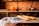 A kitchen counter displays a cloth-covered table with an assortment of baked goods including croissants, chocolate muffins dusted with powdered sugar, and several baguettes. In the background, a rustic kitchen setting features a brick wall, and various fruits sit together in a bowl beside a tall bottle.