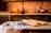 A kitchen counter displays a cloth-covered table with an assortment of baked goods including croissants, chocolate muffins dusted with powdered sugar, and several baguettes. In the background, a rustic kitchen setting features a brick wall, and various fruits sit together in a bowl beside a tall bottle.