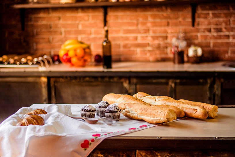 A kitchen counter displays a cloth-covered table with an assortment of baked goods including croissants, chocolate muffins dusted with powdered sugar, and several baguettes. In the background, a rustic kitchen setting features a brick wall, and various fruits sit together in a bowl beside a tall bottle.