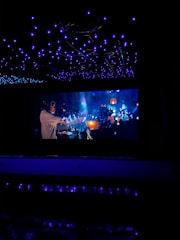 A dimly lit space with a large screen displaying a vibrant scene. The ceiling is adorned with numerous blue lights creating a starry effect. The screen depicts a busy, colorful atmosphere with people, glowing lights, and dynamic motion.