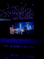 A dimly lit space with a large screen displaying a vibrant scene. The ceiling is adorned with numerous blue lights creating a starry effect. The screen depicts a busy, colorful atmosphere with people, glowing lights, and dynamic motion.