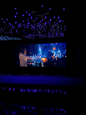 A dimly lit space with a large screen displaying a vibrant scene. The ceiling is adorned with numerous blue lights creating a starry effect. The screen depicts a busy, colorful atmosphere with people, glowing lights, and dynamic motion.