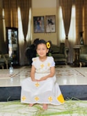 A young girl wearing a white dress with yellow designs sits on a tiled floor inside a luxurious living room. She has a sunflower in her hair and is barefoot. The room has elegant furniture, curtains, and framed photos on the wall.