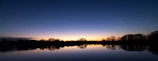 A calm lake reflecting olive green trees under a clear sky at sunset.