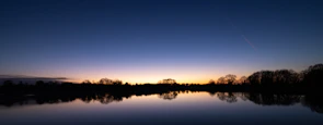 A calm lake reflecting olive green trees under a clear sky at sunset.