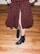 A person wearing a knee-length skirt with a black base and red floral pattern, standing on a wooden floor. The skirt has a front slit, revealing part of the leg. The person is also wearing dark blue, shiny ankle boots.