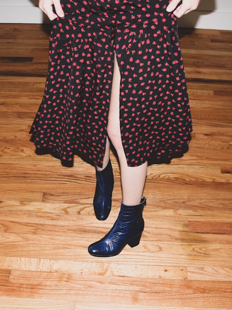 A person wearing a knee-length skirt with a black base and red floral pattern, standing on a wooden floor. The skirt has a front slit, revealing part of the leg. The person is also wearing dark blue, shiny ankle boots.