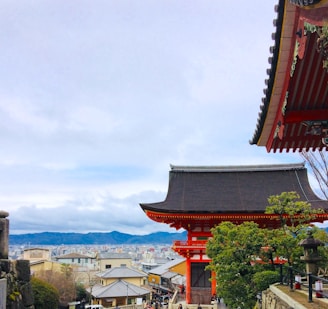A traditional Japanese temple with intricate red and black architecture stands prominently, with a panoramic view of a sprawling urban cityscape and mountains in the background. Green foliage and trees surround the temple, adding natural beauty to the setting.