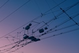 A complex network of electrical wires and cables is silhouetted against a tranquil sky with a gradient of colors ranging from soft pink to deep blue. The intricate arrangement of wires creates a pattern that is both geometric and chaotic.