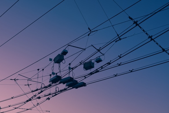 A complex network of electrical wires and cables is silhouetted against a tranquil sky with a gradient of colors ranging from soft pink to deep blue. The intricate arrangement of wires creates a pattern that is both geometric and chaotic.