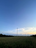 Wind turbines are situated in an expansive open field under a vast blue sky. The horizon is marked with blurred trees and bushes, creating a pastoral and serene scene. The clouds in the sky are sparse, with the sun casting a soft light, indicating either early morning or late evening.