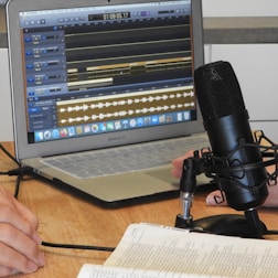 A laptop displaying audio editing software is on a wooden table. Nearby is a large microphone mounted on a stand, and the hands of two people who appear to be talking or recording. An open book is also visible, suggesting note-taking or script reading.